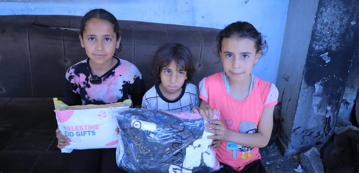 Three children sit on a couch holding packages labeled "Palestine Eid Gifts 2025" and clothing items.